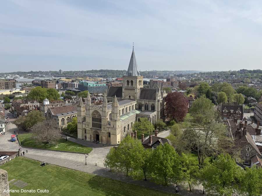 Sob um céu nublado, vê-se uma catedral bem antiga, cercada por outras construções. Há muitas árvores entre as edificações. O ângulo da visão parte de cima, talvez da altura da janela de um edifício.