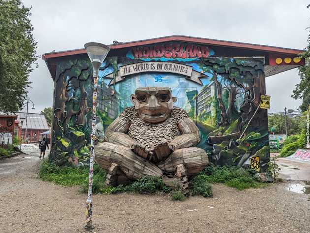 'Em frente a um mural colorido com escritos como "Wonderland" e "The world is in our hands", vemos uma estrutura de ripas de madeira na forma de um gigante sentado. Seus olhos são representados por dois furos.'