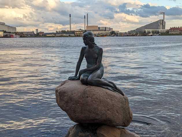 'Em um grande canal, vemos em primeiro plano a escultura de uma sereia sentada sobre uma pedra, observando o horizonte. Ao fundo, prédios de diferentes alturas com longas chaminés.'